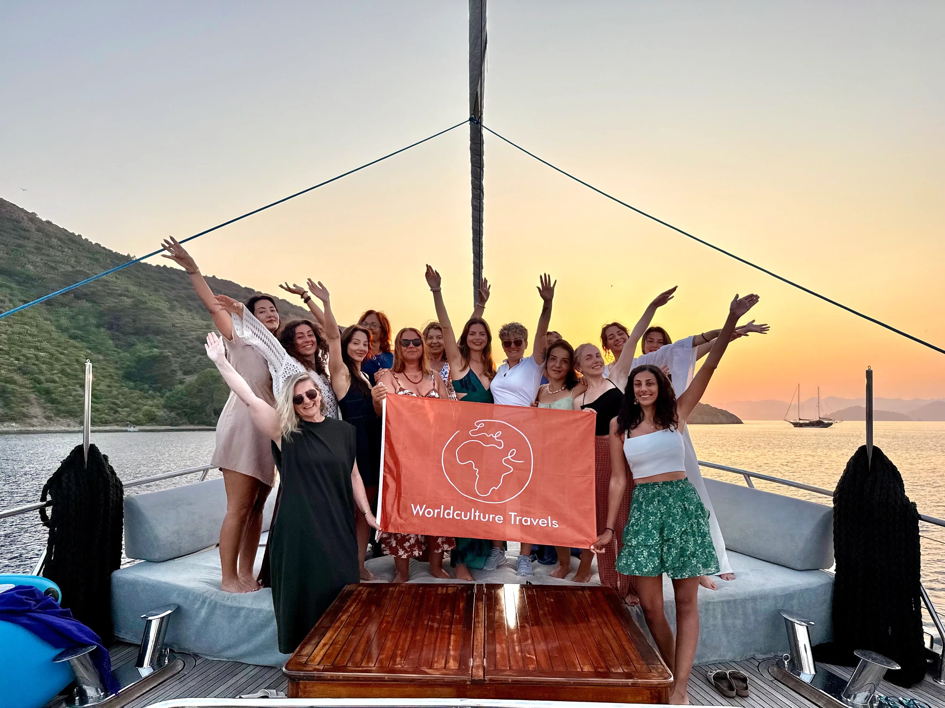 Die Gruppe jubelt mit erhobenen Armen und der Worldculture-Travels-Flagge auf dem Bug im Sonnenuntergang.