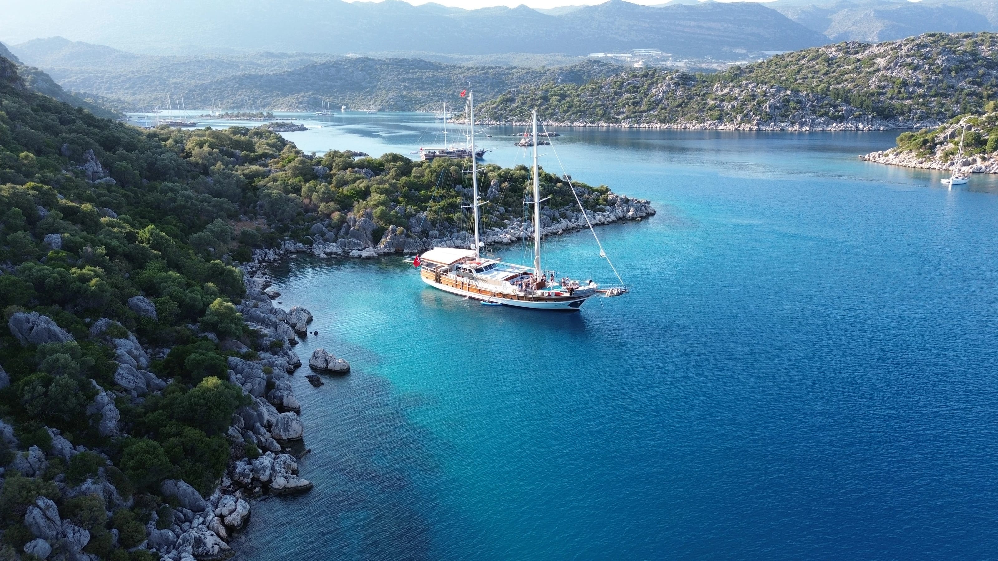 Aus der Luft gesehen liegt die Yacht in einer türkisfarbenen Bucht zwischen felsigen, bewaldeten Ufern.