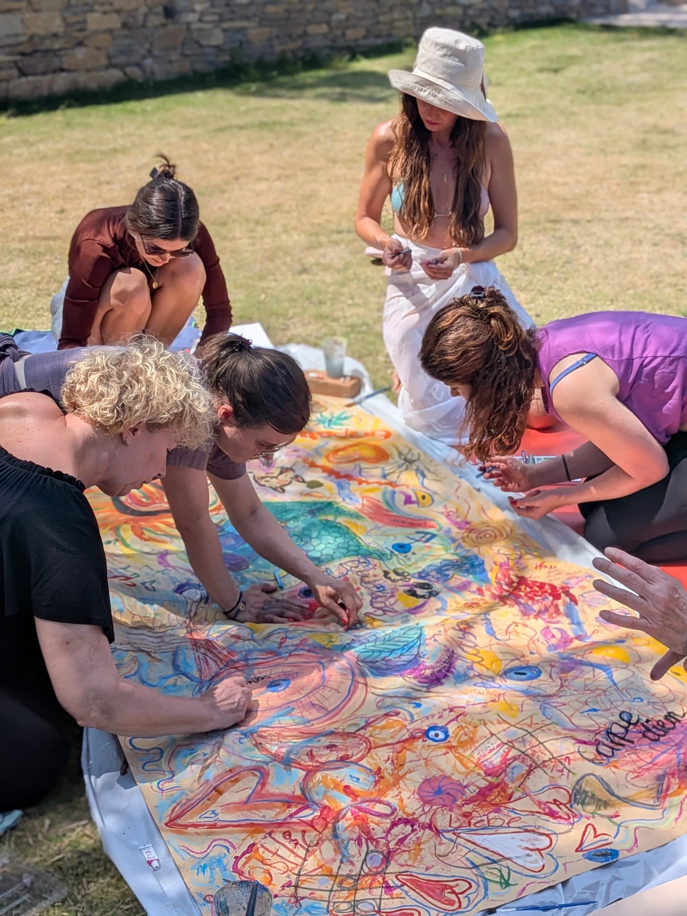 Mehrere Frauen knien im Gras und malen gemeinsam an einem großen, bunten Bild.